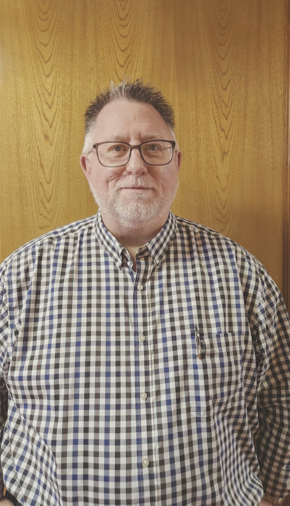 A middle-aged man with short hair, glasses, and a trimmed beard stands in front of a wooden wall. He is wearing a black, blue, and white checkered shirt with a pen in the pocket.